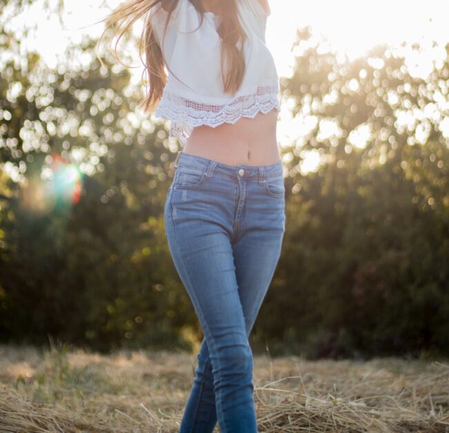 Woman Wearing White Crop Top in Blue Jeans in Field with sun behind her.