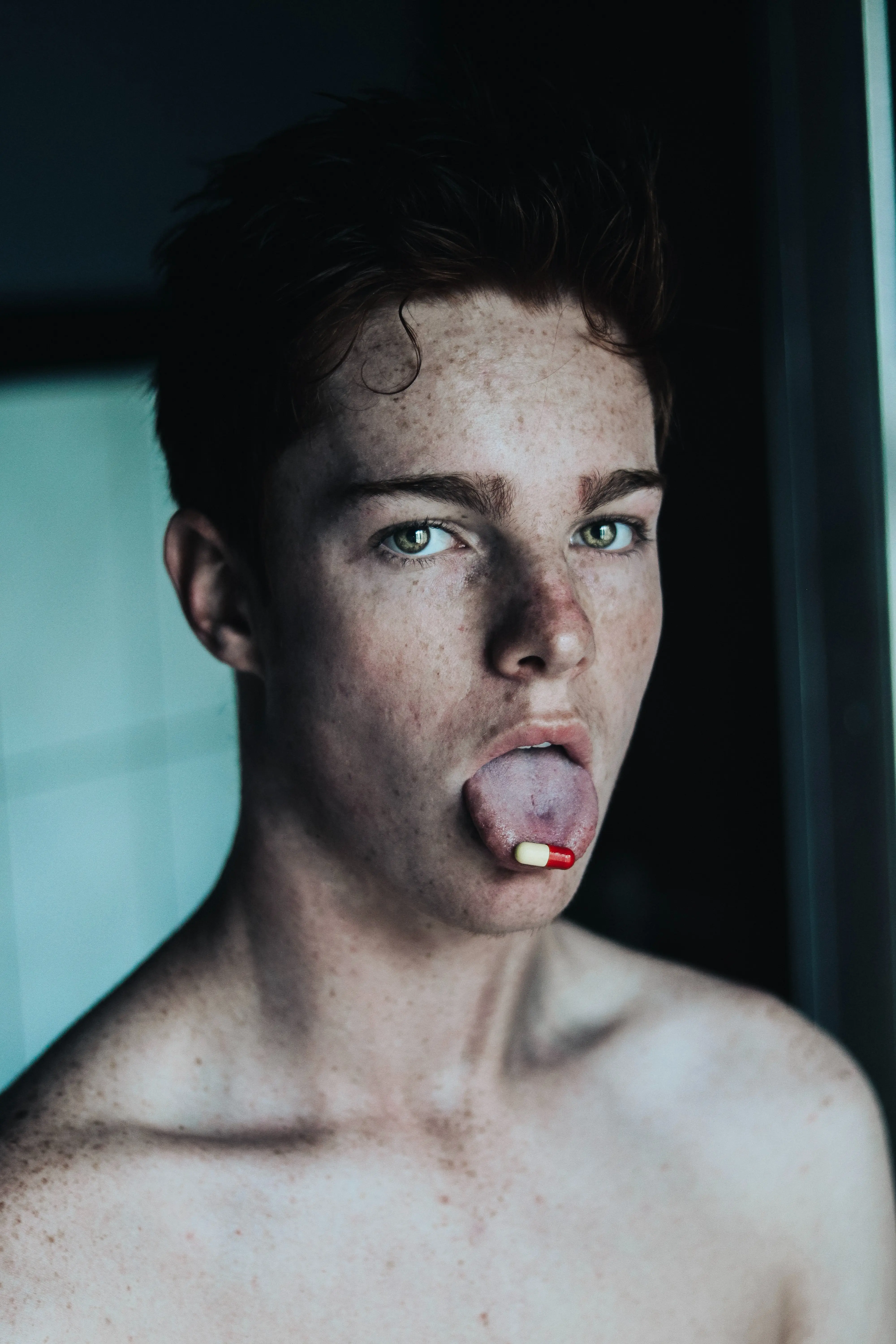Topless man with a red and white pill on the tip of his tongue.