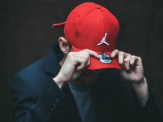 Man Pulling Red Air Jordan Hat Down Over His Eyes Wearing a Black Jacket and Grey Shirt.