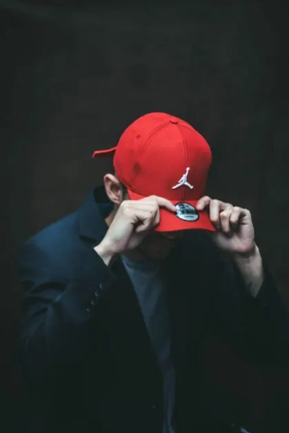 Man Pulling Red Air Jordan Hat Down Over His Eyes Wearing a Black Jacket and Grey Shirt.