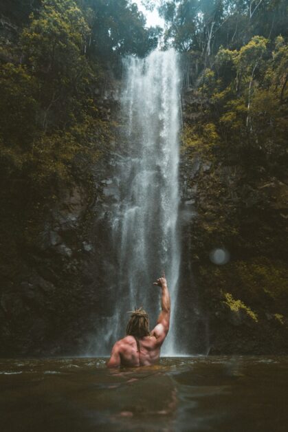 Man Standing Facing Waterfall in Water with Shirt Off