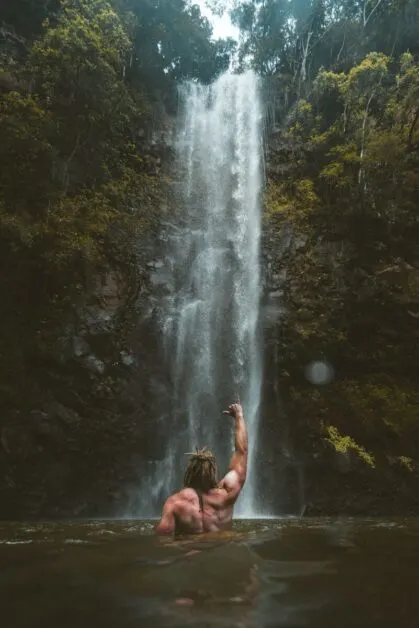 Man Standing Facing Waterfall in Water with Shirt Off
