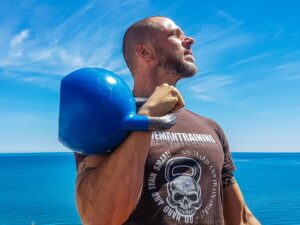 Guy Holding Blue Kettlebell With Ocean Behind