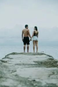 Shirtless Man and Woman in Bathing Suit Top and Jean Shorts Standing On Rock Overlooking Water