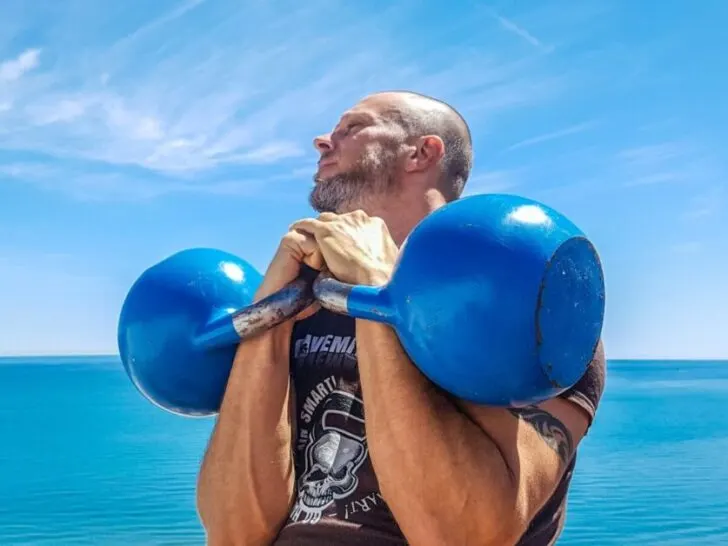 Guy Holding Two Blue Kettlebells with Ocean in Background