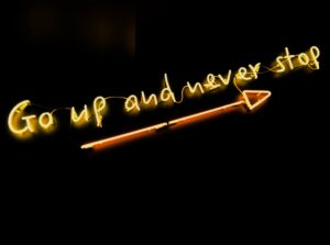 Neon sign that says go up and never stop with arrow pointing upwards underneath the words