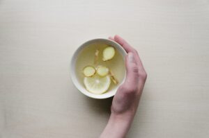 Woman's hand holding white cup with ginger and lemon in hot water.