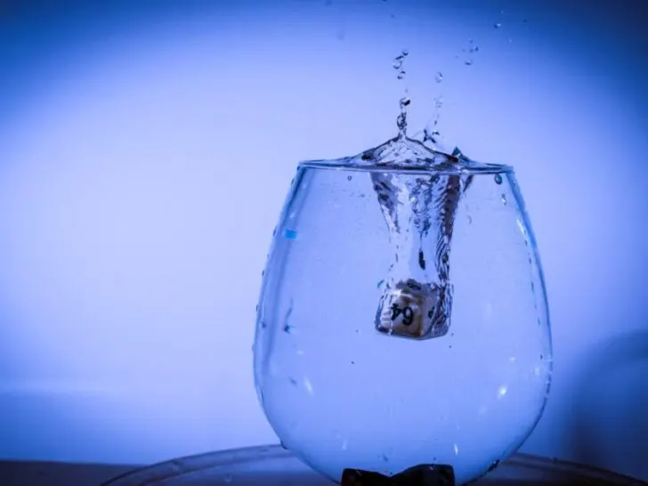 Glass of water with blue background and a cube of ice dropping in.