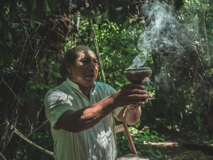 Mayan medicine man in jungle holding cup with smoke floating into air.